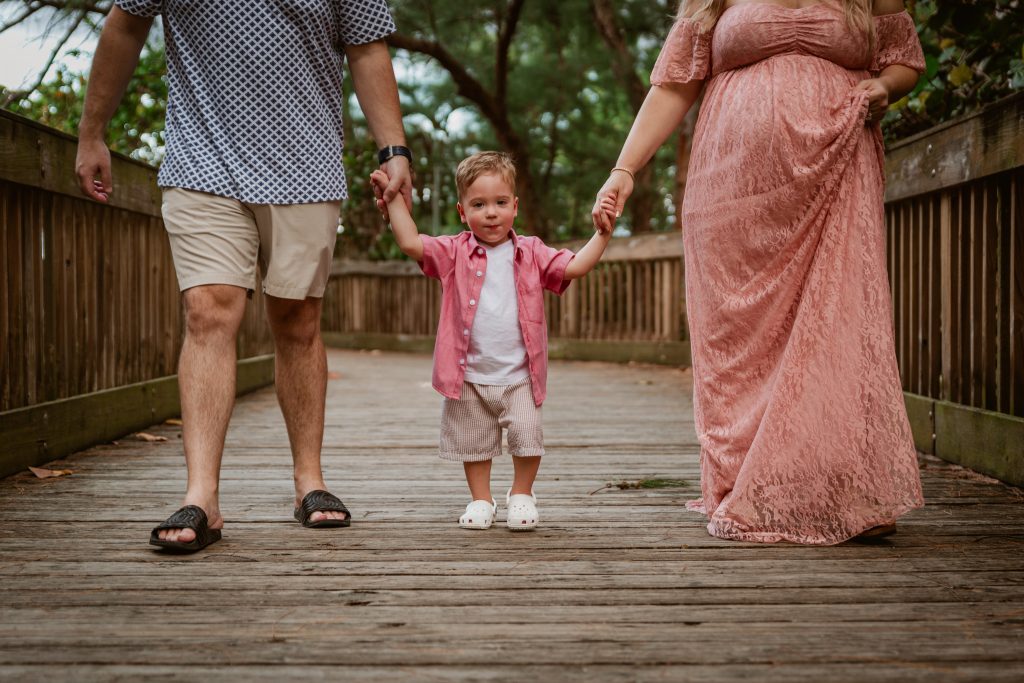 Pregnant couple with a toddler on a boardwalk at the beach.